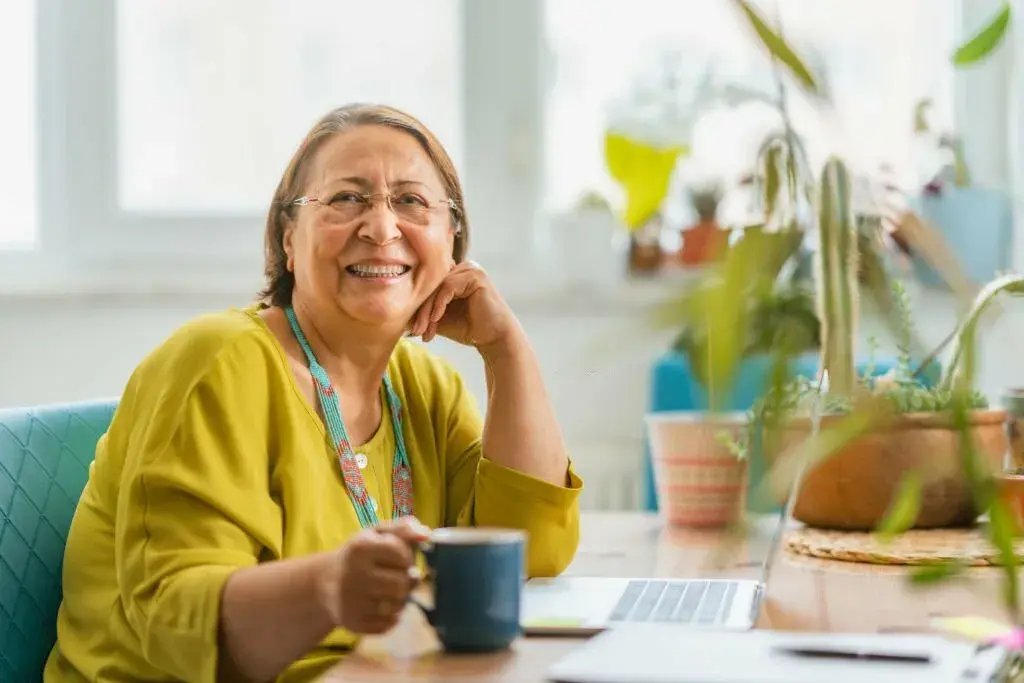 Senior woman smiling at the camera with a cup of coffee and a laptop, in a bright home setting