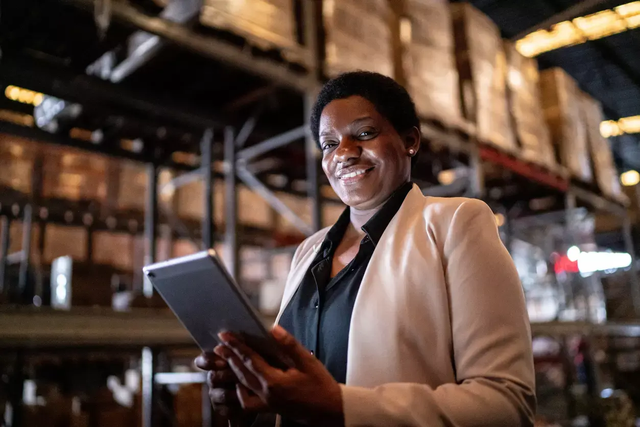 A confident businesswoman in a warehouse environment, holding a tablet and smiling.
