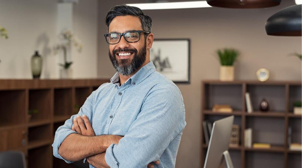 Hombre sonriente con brazos cruzados en una oficina, reflejando confianza y profesionalismo en el ámbito laboral